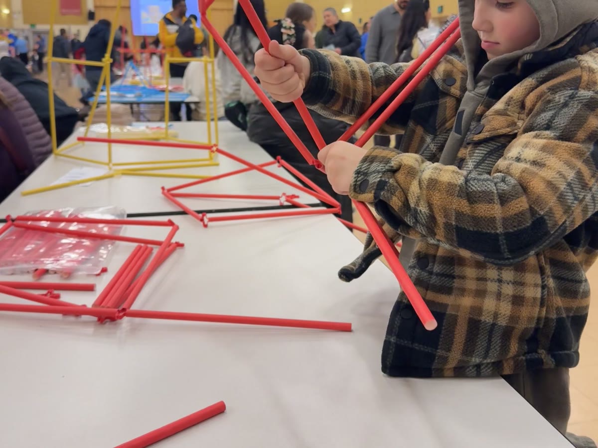 A young builder connects red straws into a Straw Structure at a table covered in parts